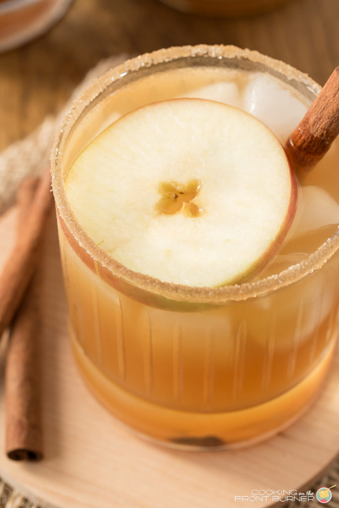 a short cocktail glass with apple pie cinnamon cocktail with an apple floating on top and cinnamon stick on side