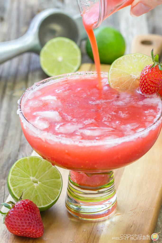 a margarita glass with a strawberry margarita being poured in with a lime and strawberry on glass