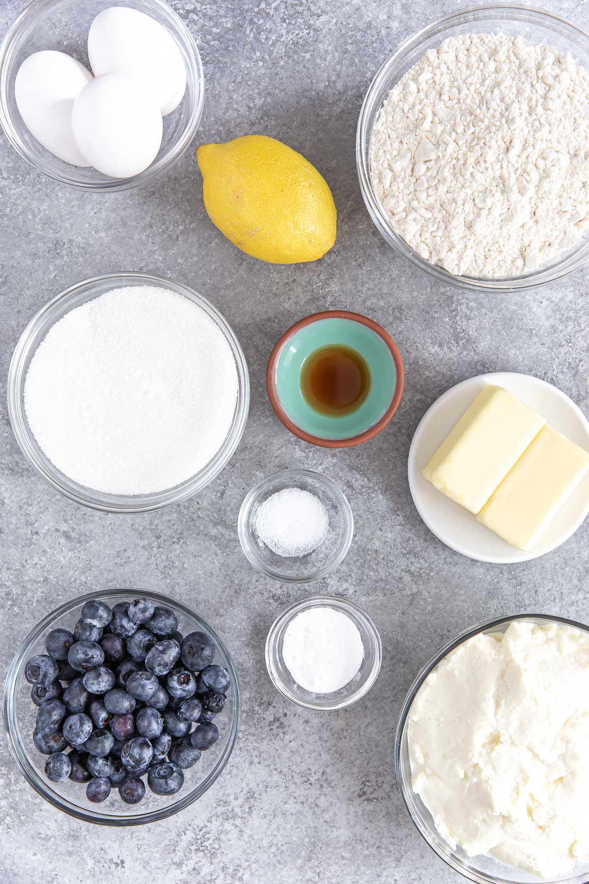 flour, sugar, lemon, vanilla extract, eggs, blueberries ricotta cheese and butter in bowls on a counter