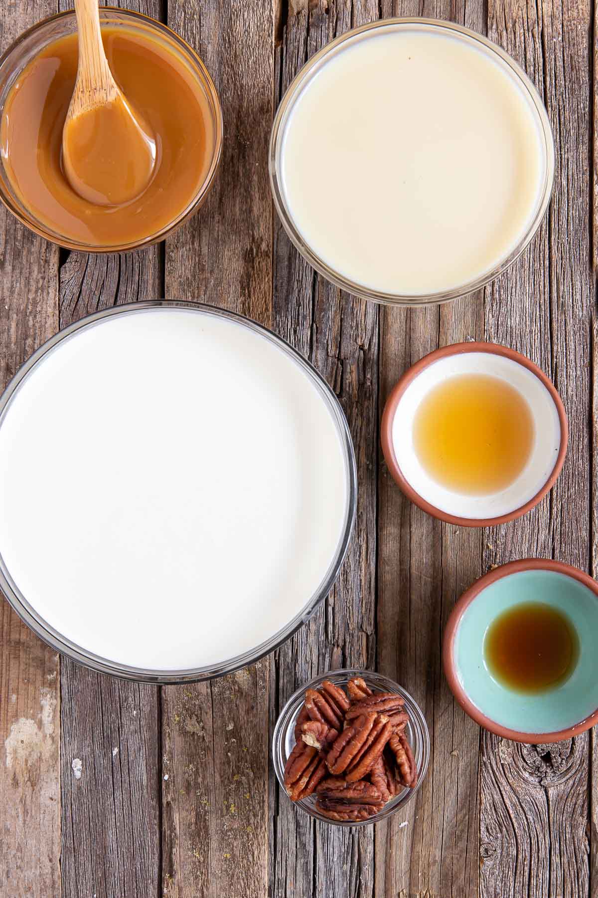 bowls of ingredients needed to make bourbon pecan caramel ice cream - whipped cream, caramel sauce, condensed milk, pecans, bourbon and vanilla extract