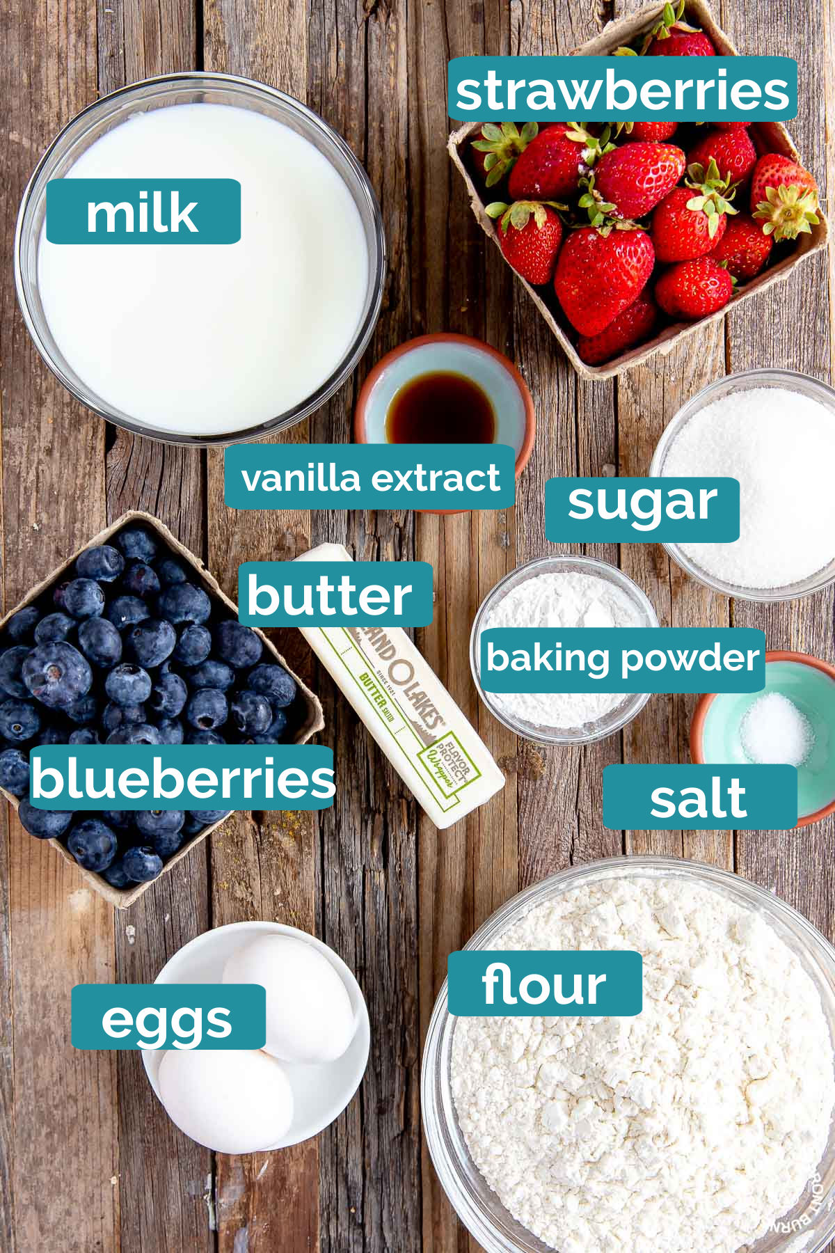 milk, strawberries, vanilla extract, blueberries, sugar, baking powder, salt, eggs and flour on a board in bowls