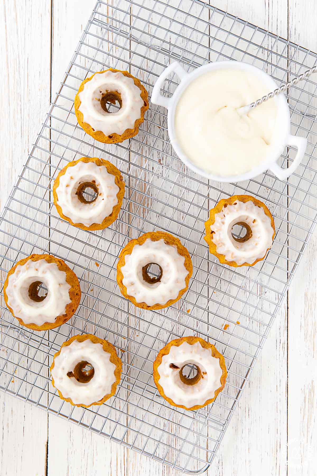 7 mini pumpkin spice cakes on a rack with a bowl of frosting