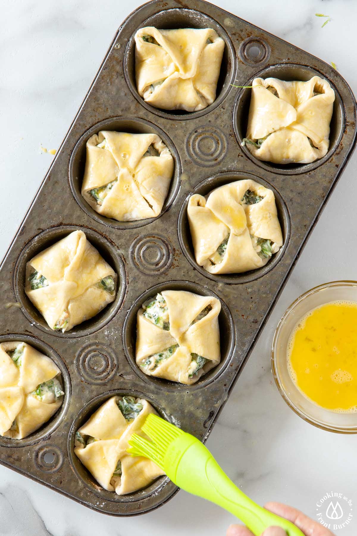 muffin tin with unbacked spinach puffs brushing egg wash on top