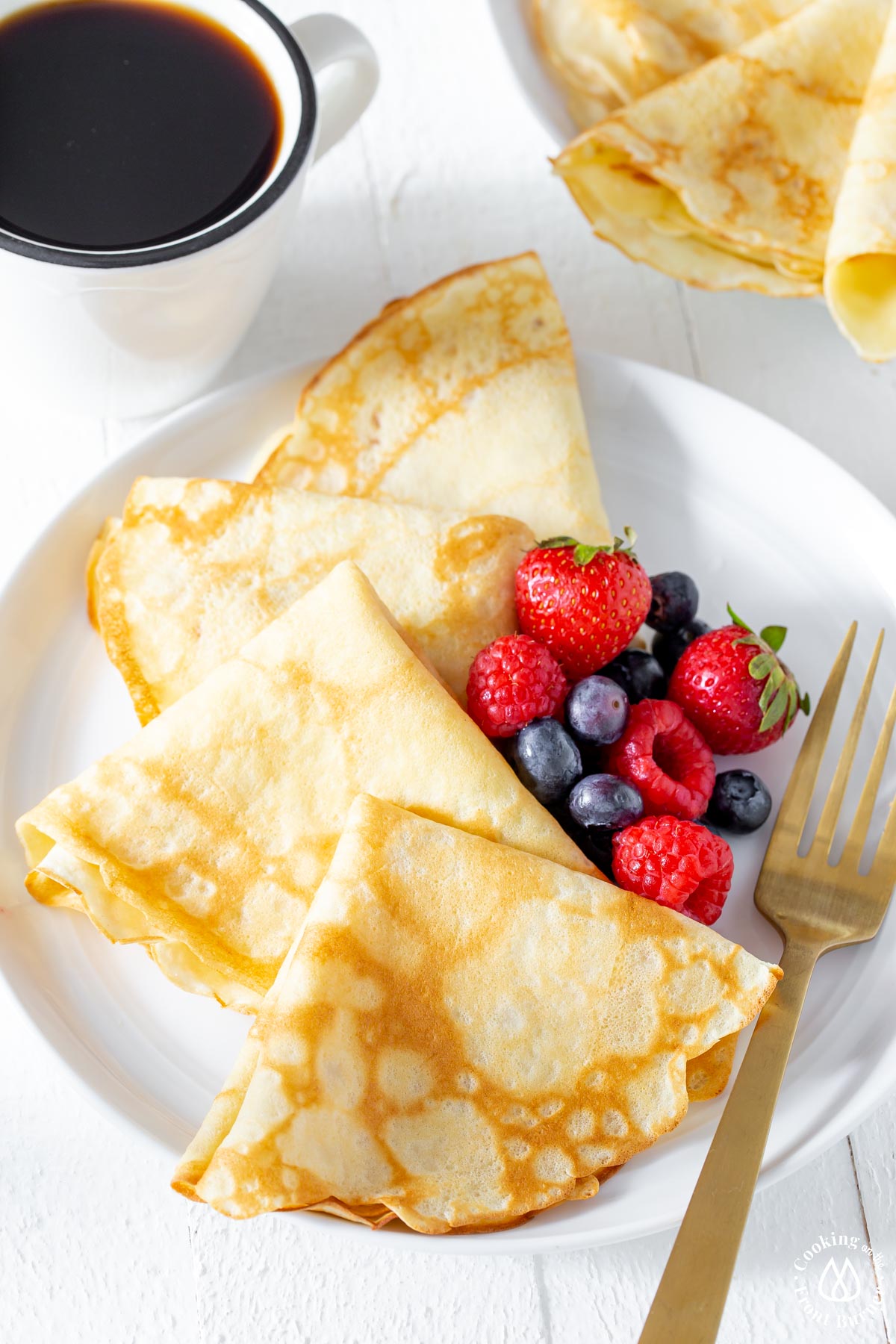 white plate with 4 crepes and a side of berries with a coffee cup